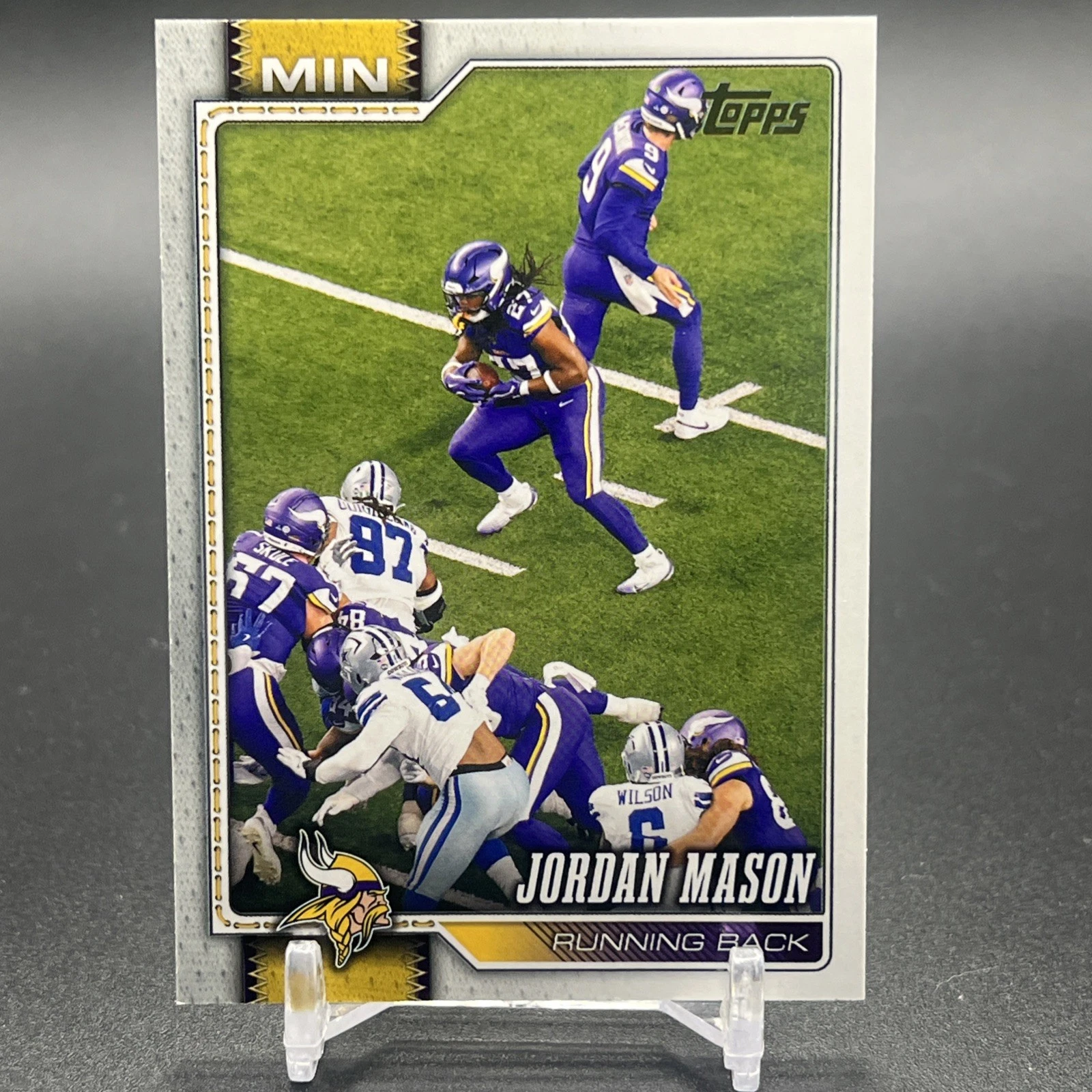 Actual independent photograph of Jordan Mason 2026 Topps Flagship Golden Mirror #130, carrying the ball in a purple Minnesota uniform with Dallas defenders in the foreground. This reference copy is shown on a clear stand and is not the submitted card.